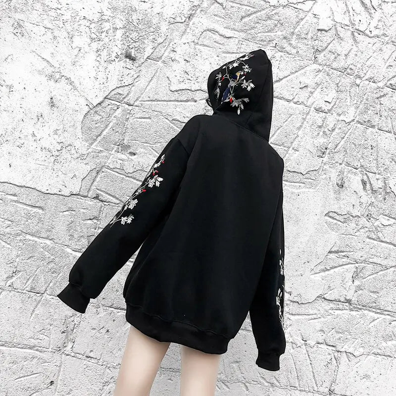 Person wearing a black hoodie with floral embroidery against a textured gray wall.