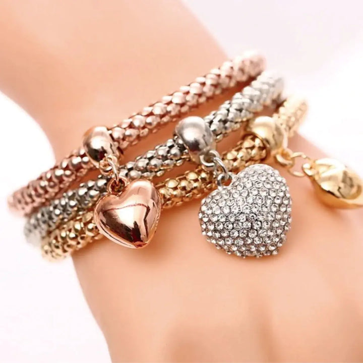 Stack of gold, silver, and rose gold bracelets with heart charms on a wrist.